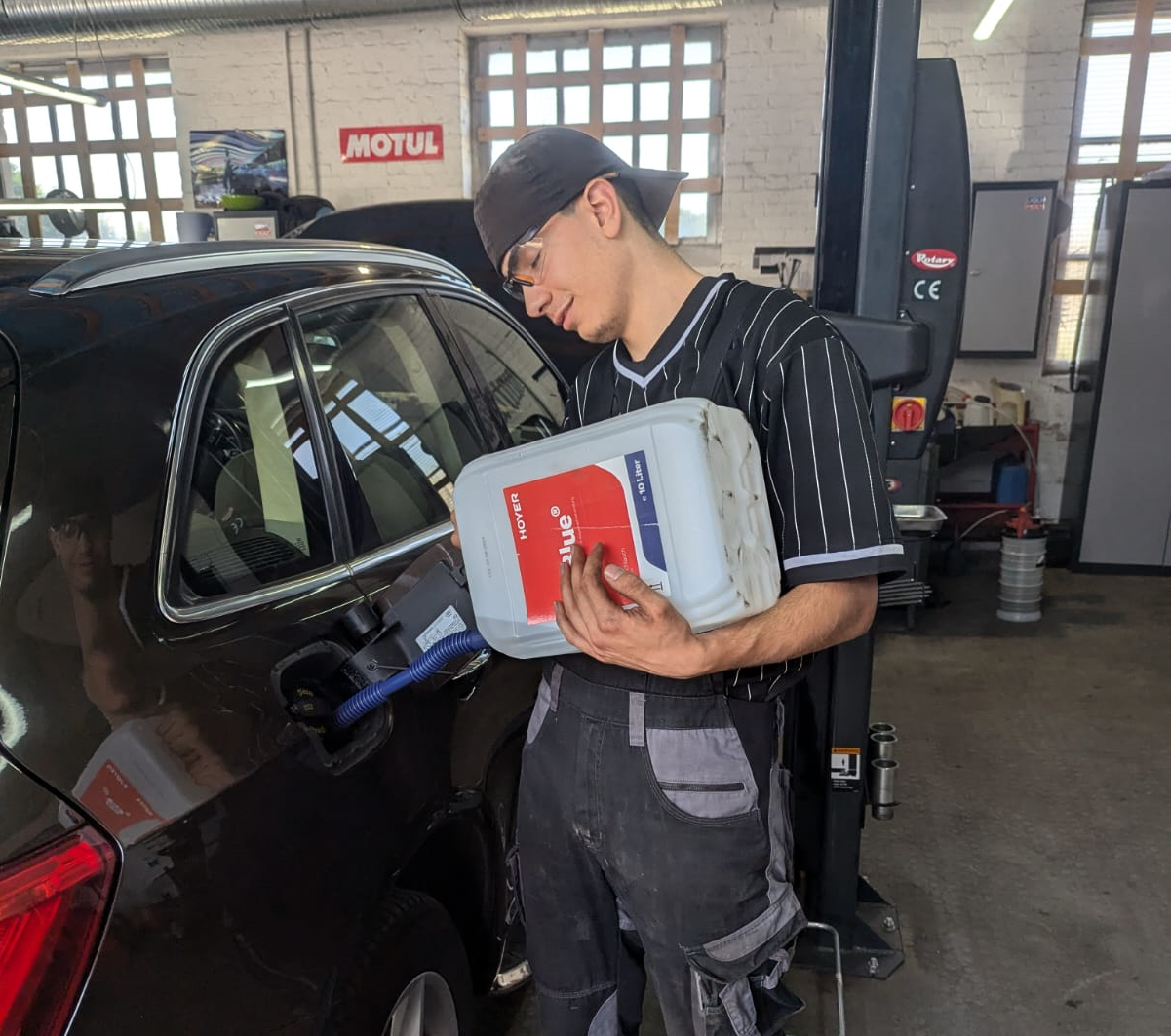 Ein junger Mann in Arbeitskleidung steht neben einem Auto in einer KFZ-Werkstatt und füllt Flüssigkeit in den Tank eines Autos.