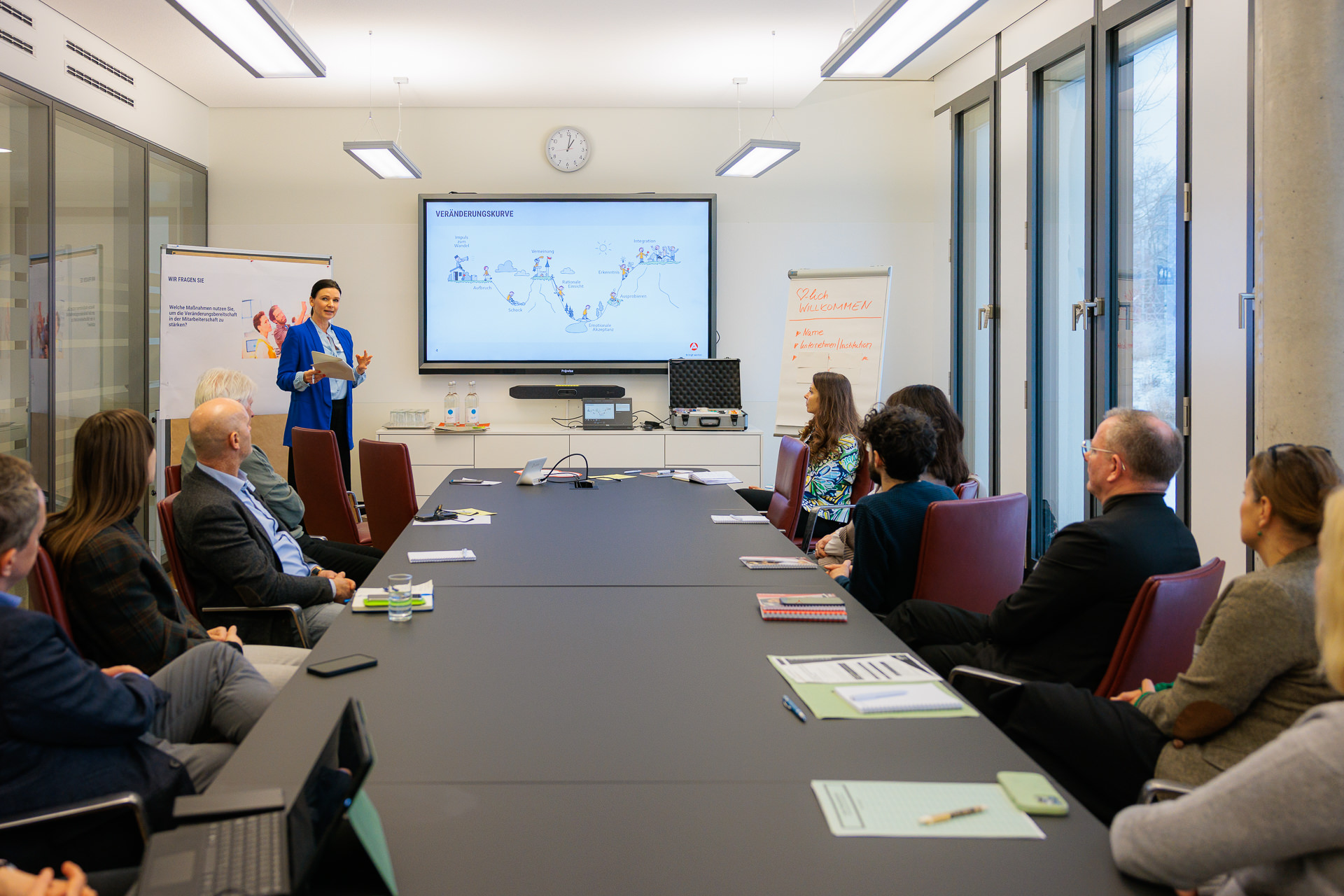 Bild: Länglicher Konferenzsaal, am Tisch rechts und links Teilnehmende, ganz hinten Bianca Naue von der Agentur für Arbeit Potsdam.