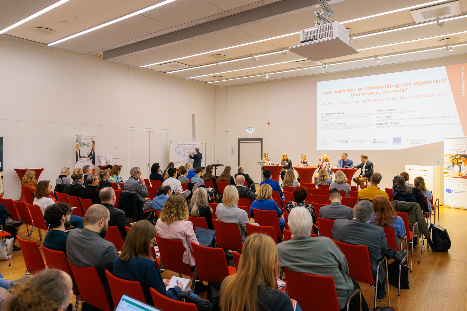 Bild: Voll besetzte Stuhlreihen, im Hintergrund die Podiumsdiskussion auf der Bühne, wo gerade Dr. Friederike Haase spricht.