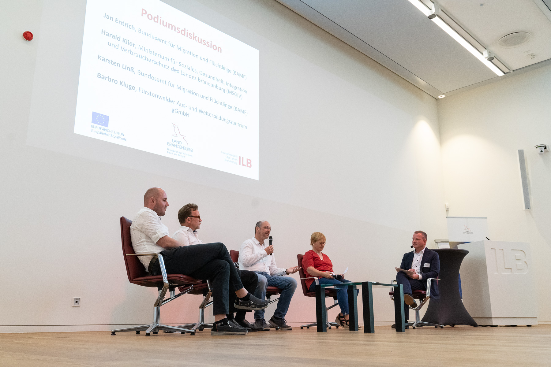Bild: Bild zeigt Podiumsdiskussion, aufgenommen links neben der Bühne. Auf dem Podium von links: Jan Entrich (BAMF), Harald Klier (MSGIV), Karsten Linß (BAMF), Barbro Kluge (FAWZ) und Dr. Matthias Kirbach (Moderator)..