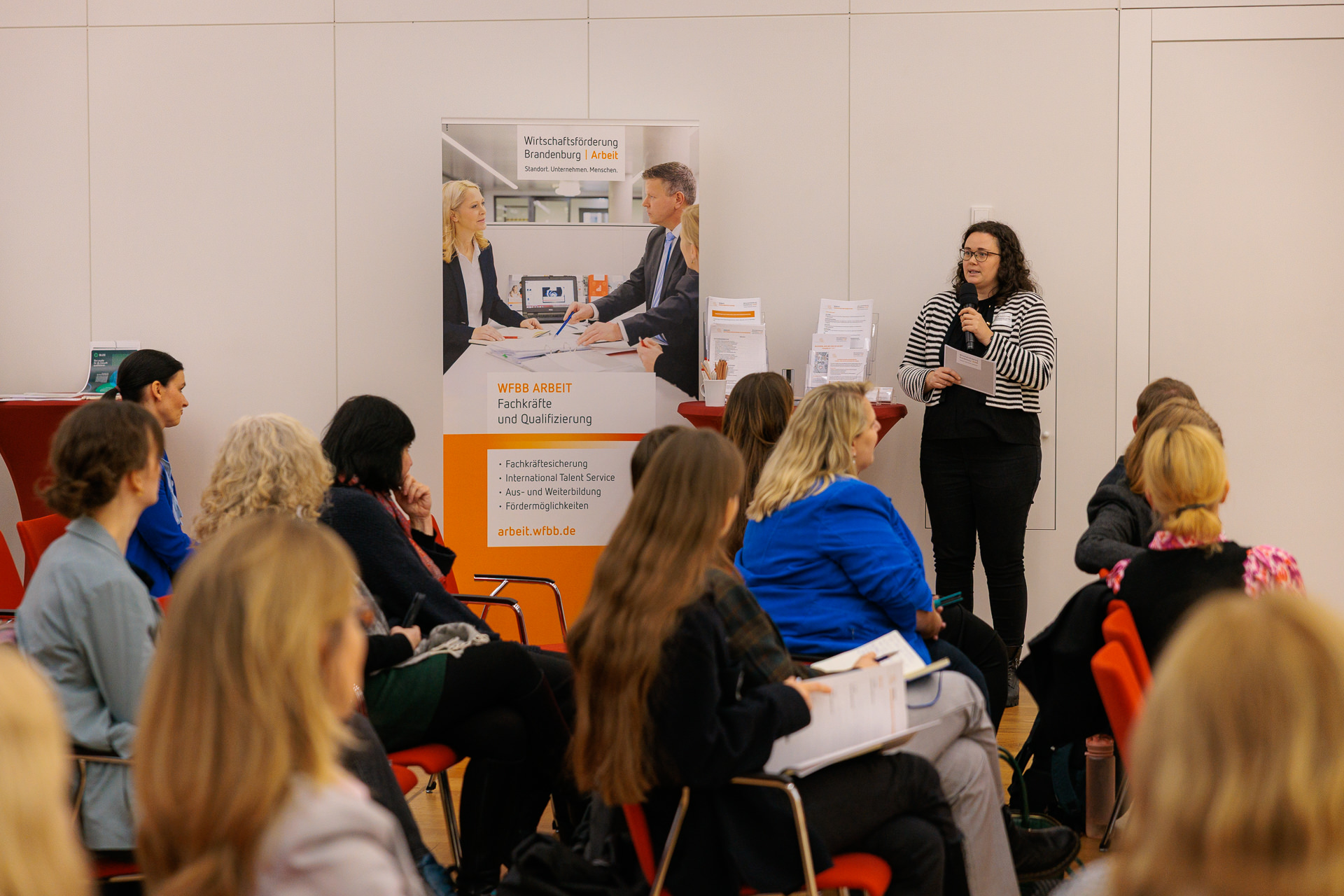 Bild: Patrizia Zimmermann von der der Wirtschaftsförderung Land Brandenburg GmbH steht neben einem RollUp an einem Stehtisch, im Vordergrund Publikum.