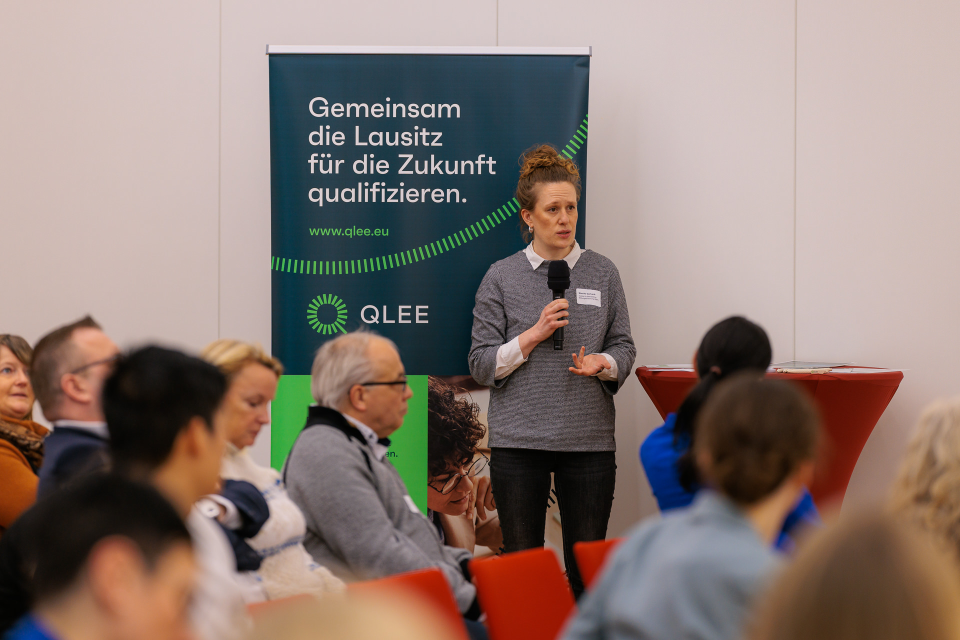 Bild: Mareike Gerhardt vom Qualifizierungsverbund für erneuerbare Energien in der Lausitz (QLEE) neben einem RollUp am Stehtisch im Vordergrund Publikum.