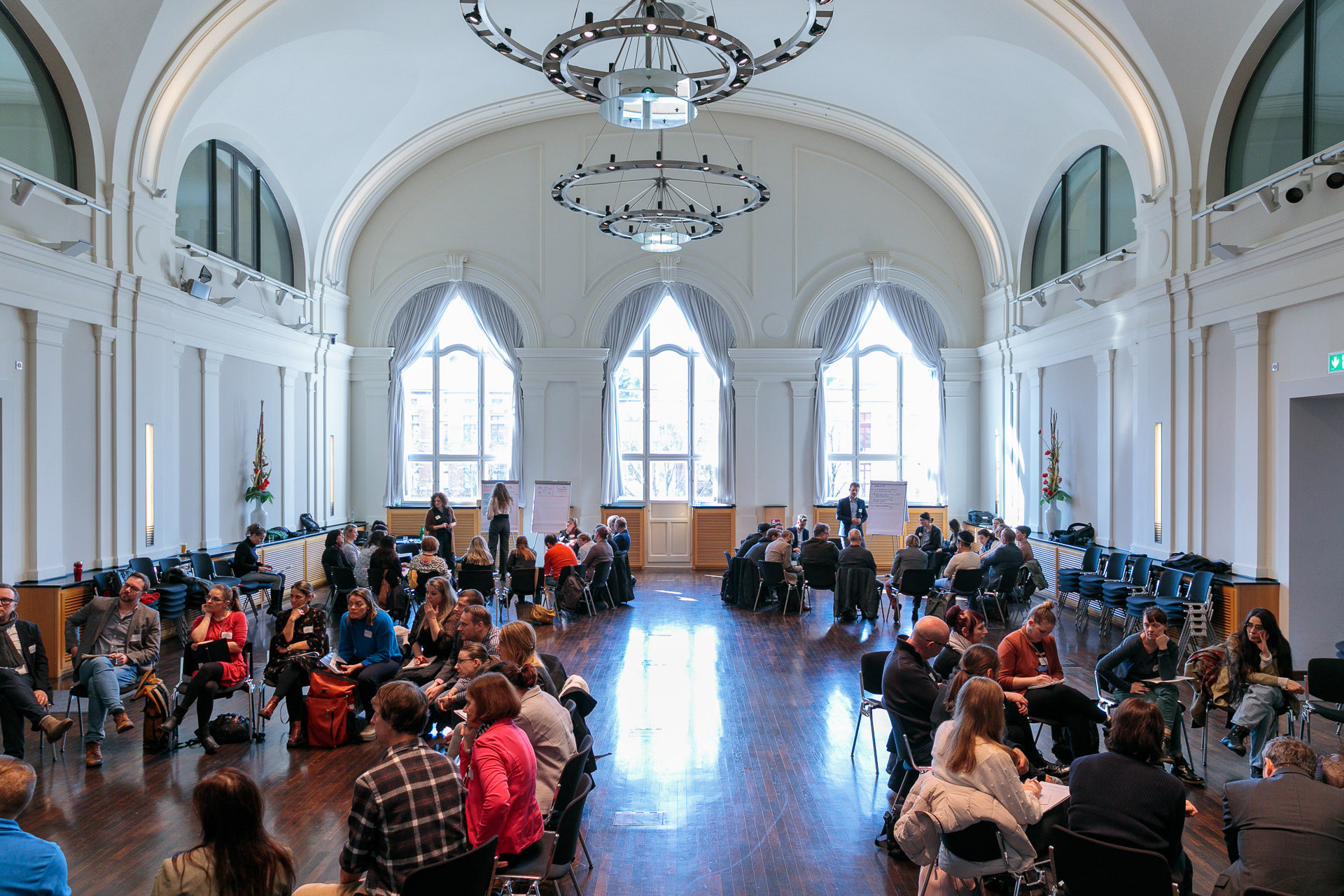 Bild: Im Brandenburg Saal der Staatskanzlei sitzen Teilnehmende im Kreis, im Hintergrund Flipcharts und riesige Fenster, die den Saal mit Tageslicht fluten.