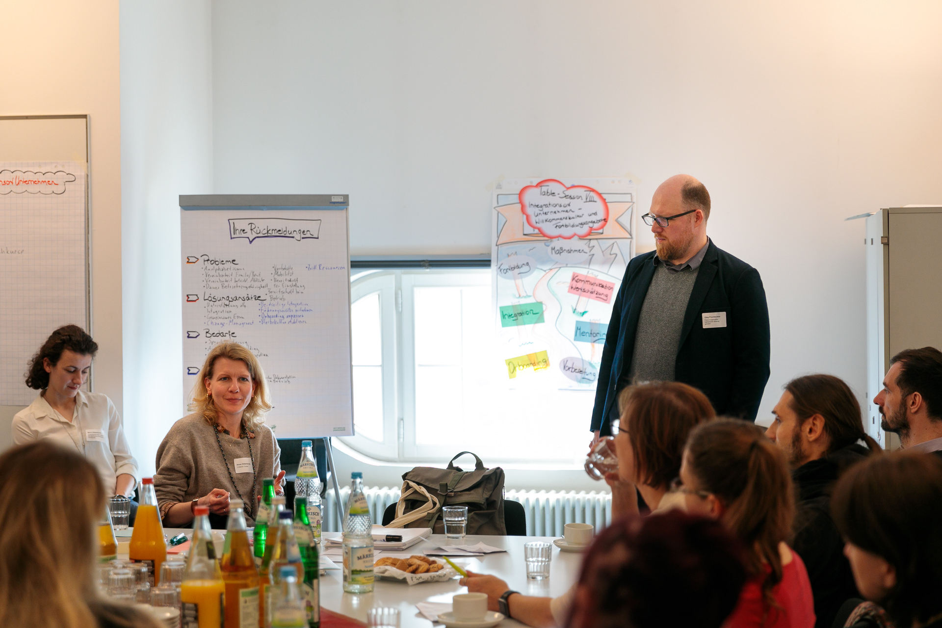 Bild: Bei Getränken und Gebäck leiten Oleg Pronitschew und Birgit Lisewitzki ein Table-Session; im Hintergrund zwei Flipcharts mit Lösungsansätzen und Diagrammen.