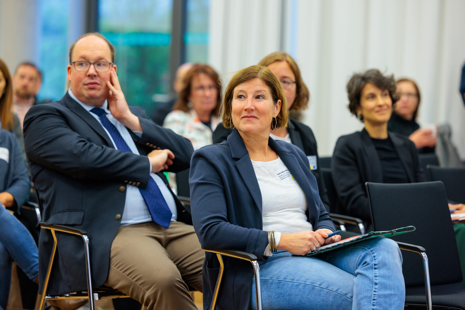 Bild: Viele Menschen sitzen in mehreren Stuhlreihen und schauen nach rechts.