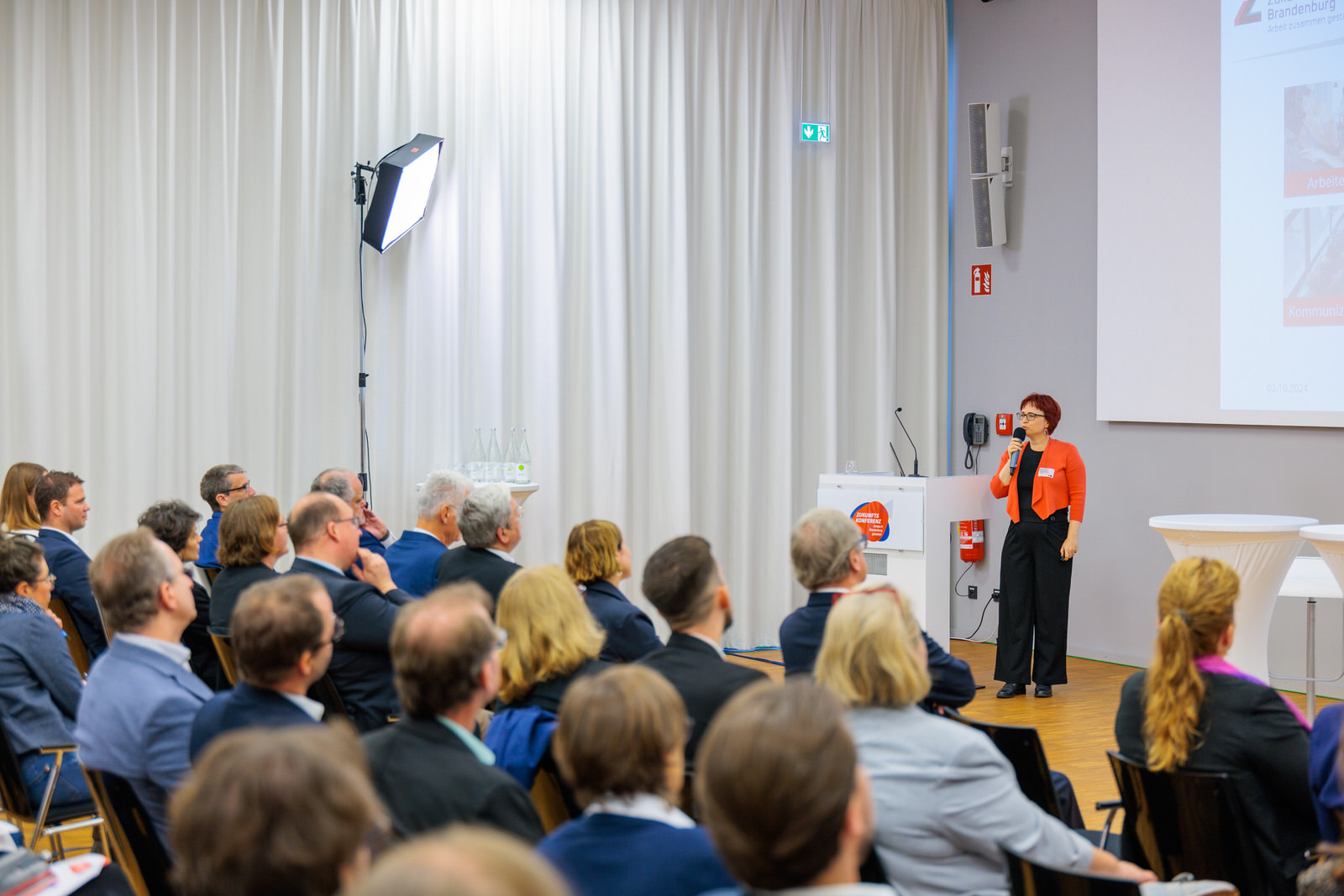 Bild: Publikum von hinten fotografiert, ganz vorne steht Denise Gramß vom Zukunftszentrum Brandenburg neben dem Rednerpult mit einem Mikro in der Hand.