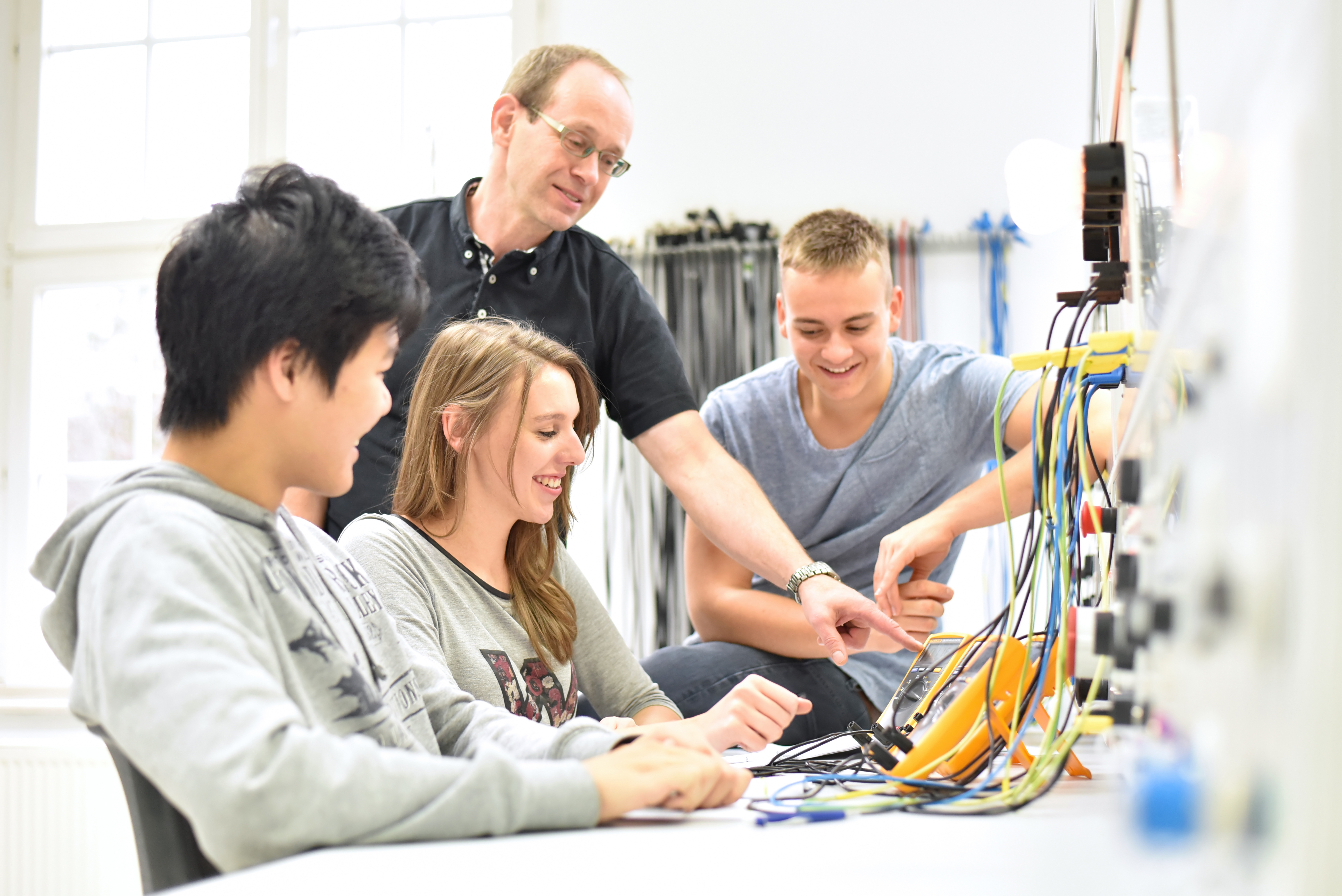 Junge Menschen sitzen vor einer technischen Einrichtung aus der viele Kabel hängen und werden von einem Ausbilder angeleitet.