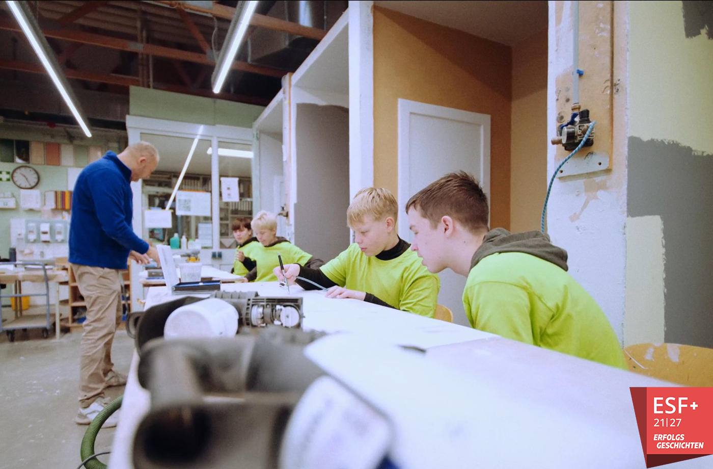 Vier Schüler sitzen in einer Werkstatt an einer Werkbank und werden bei der Arbeit vom Ausbilder auf der anderen Seite der Werkbank angeleitet.