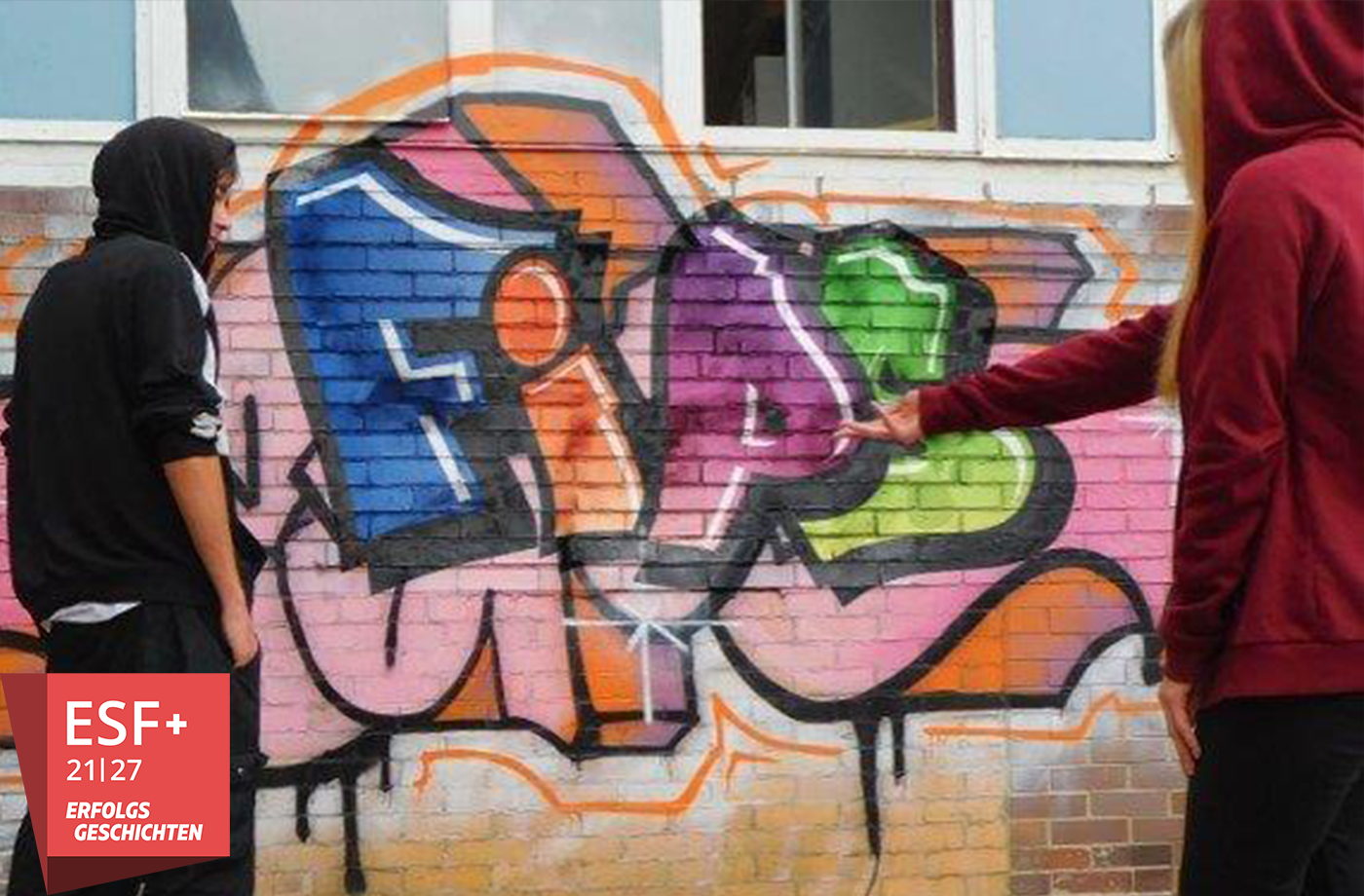 Zwei junge Menschen stehen vor einer Graffiti-Wand auf die das Wort 'FiPS' gesprüht wurde.
