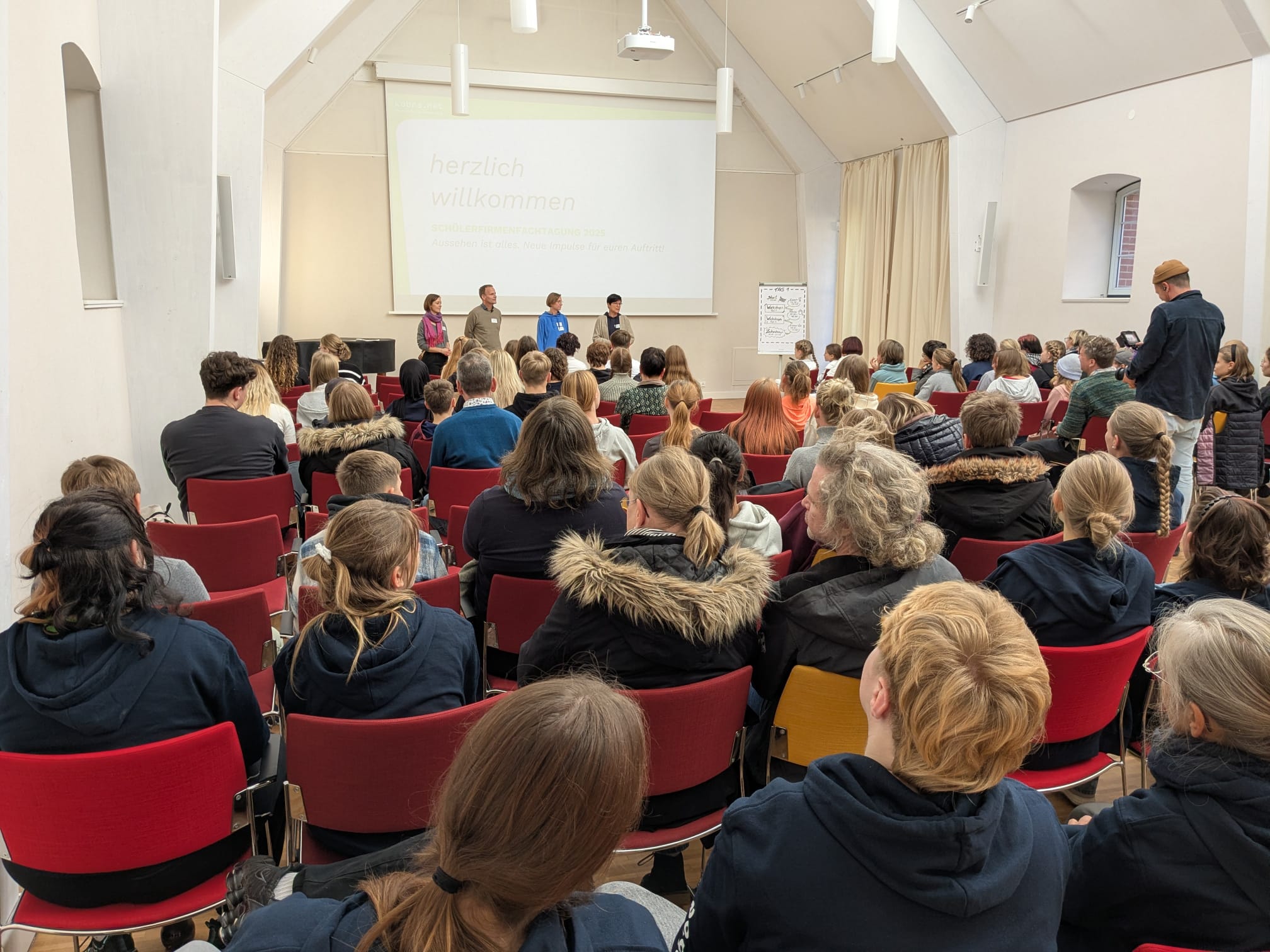 Vollbesetzte Stuhlreihen und im Hintergrund Menschen, die vor dem Publikum sprechen.