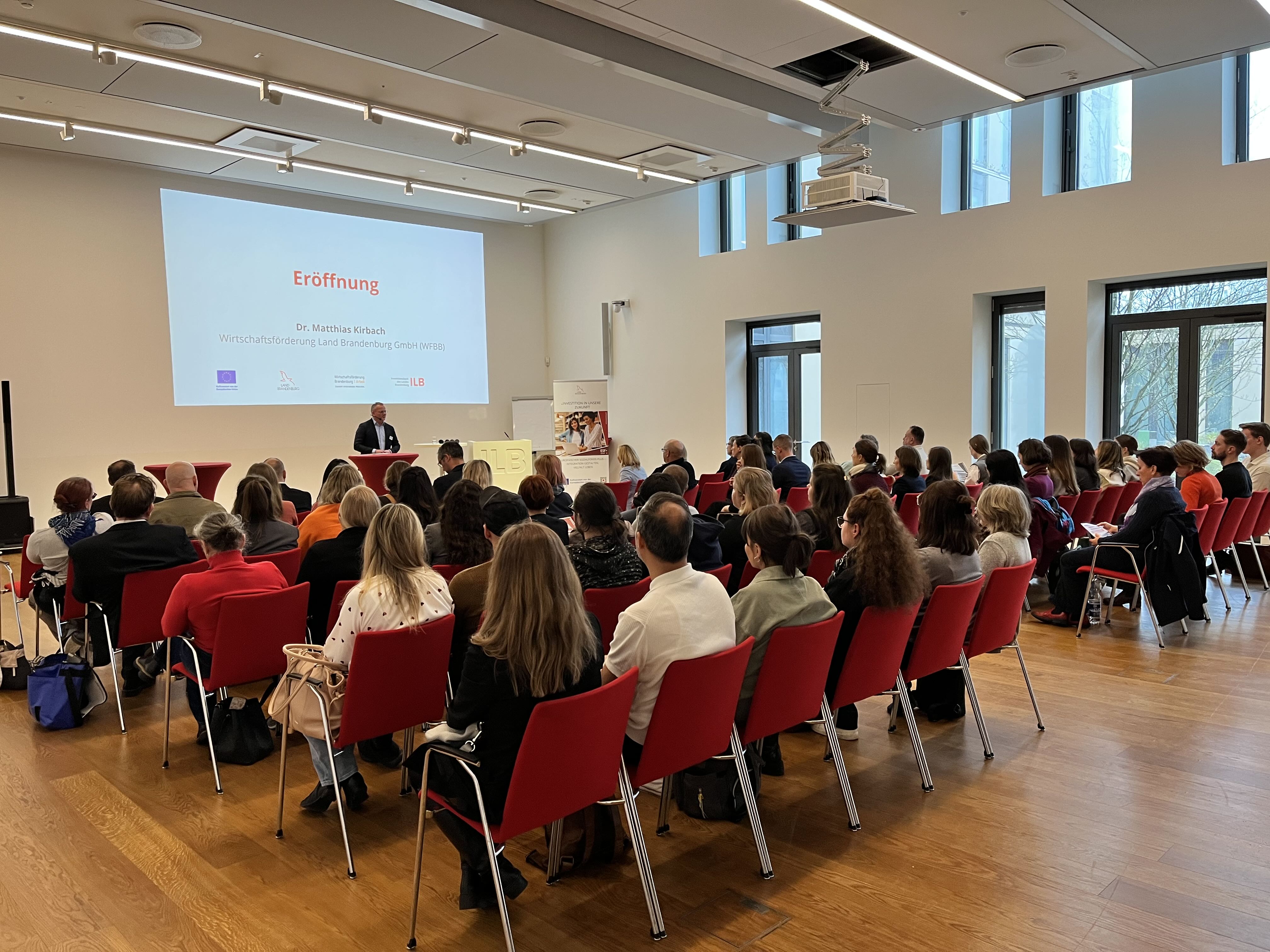 Großer Veranstaltungssaal mit voll besetzten Stuhlreihen, im Hintergrund eröffnet Moderator Dr. Matthias Kirbach die Veranstaltung.