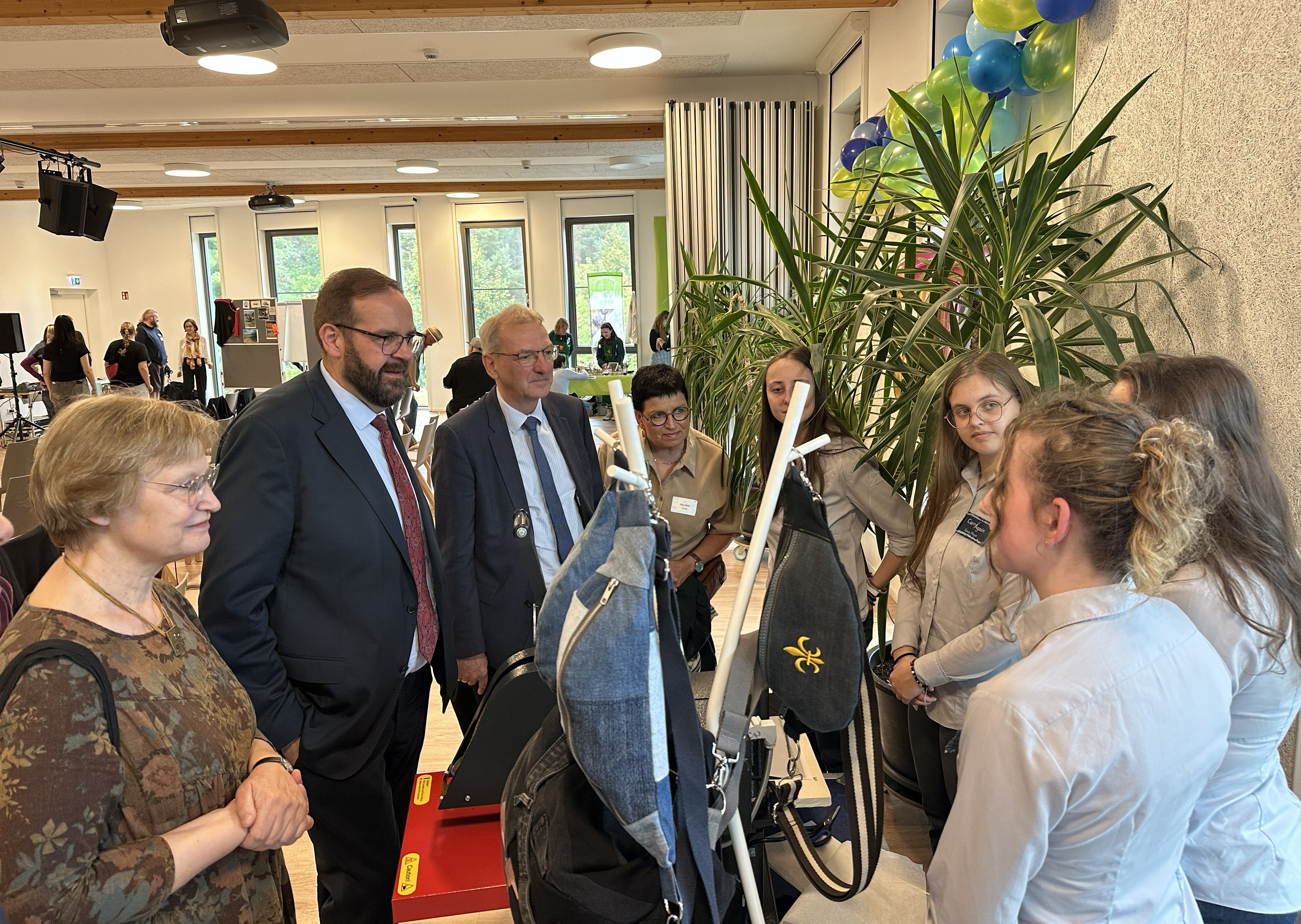 Minister Gordon Hoffmann und Staatssekretär Hendrik Fischer stehen an einem Tisch mit dort ausgestellten Taschen und sprechen mit Schülerinnen.