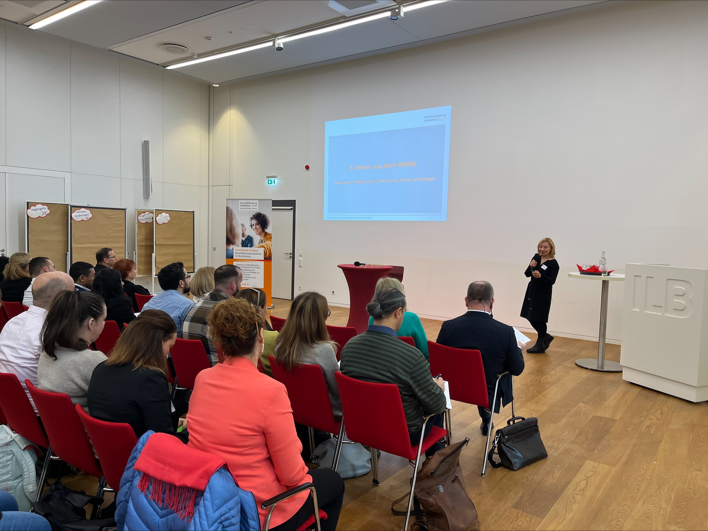 Voll besetzte Stuhlreihen. Vorne neben dem Rednerpult steht MWAE-referentin Dina Ulrich mit einem Mikro in der Hand. Im Hintergrund die Startfolie einer Präsentation auf der 'Neues aus dem MWAE' steht.