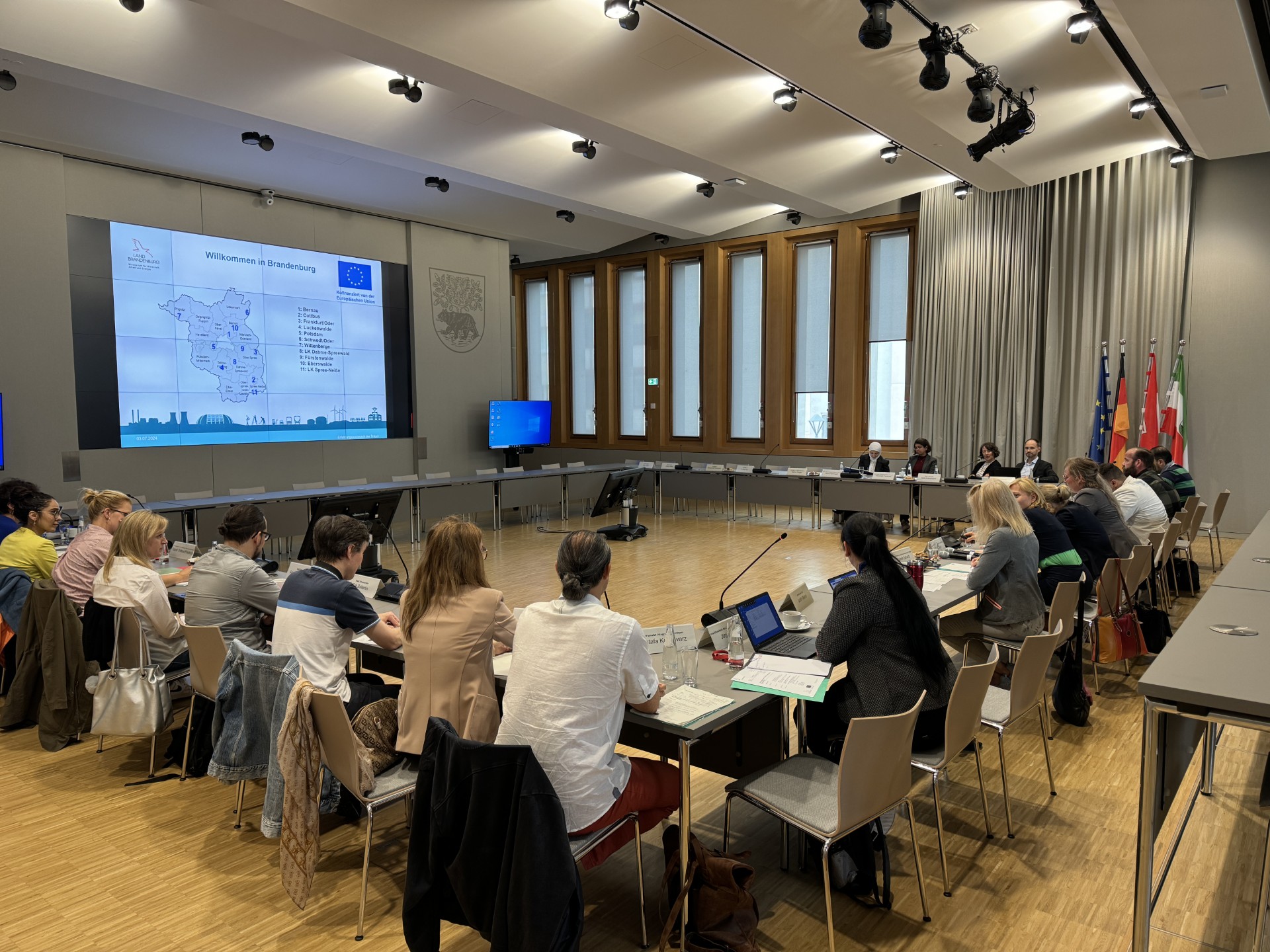 Menschen sitzen u-förmig platziert an Tischen in einem großen Saal. Im Hintergrund ist eine Präsentation mit einer Landkarte zu sehen.