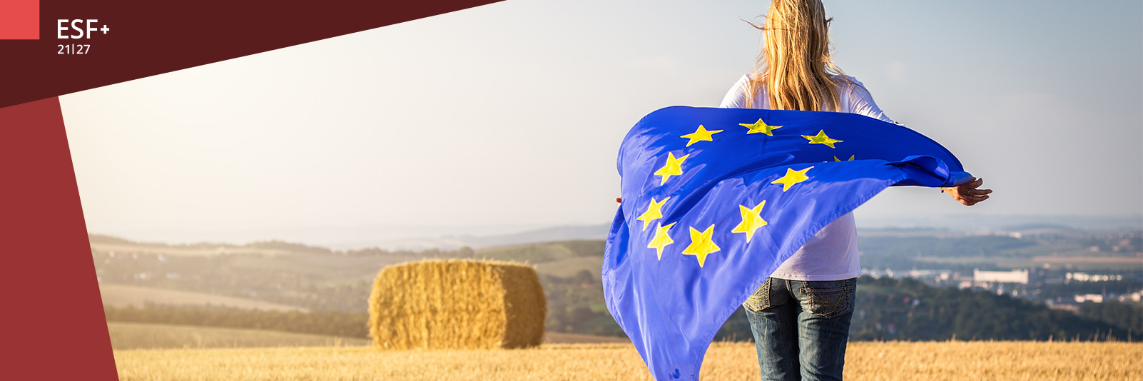 Frau steht auf einem abgeernteten Feld mit Strohballen im Hintergrund und hält eine EU-Fahne hinter ihrem Rücken, die im Wind weht.