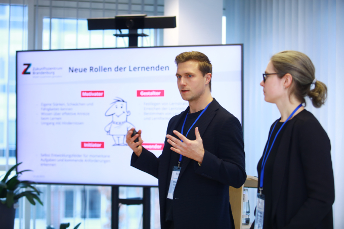 Sebastian Richter (links) und Veronika Schneider (rechts) stehen vor einem Monitor, auf dem eine Präsentation zu sehen ist und stellen diese gestikulierend vor.