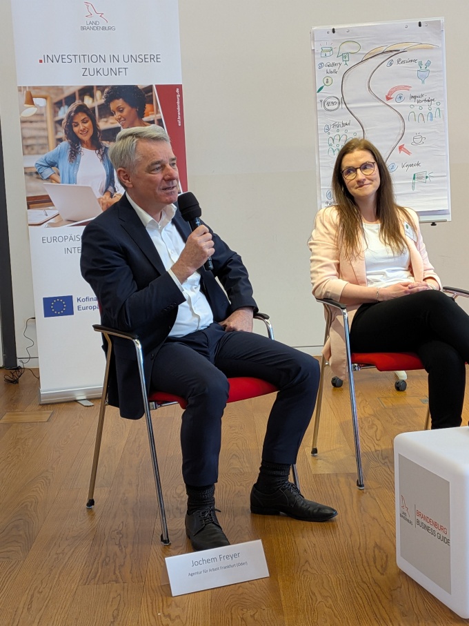 Jochem Freyer von der Agentur für Arbeit Frankfurt (Oder) sitzt auf einem Stuhl mit einem Mikro in der Hand, auf dem Stuhl daneben sitzt Jenny Schüler von der LEAG.