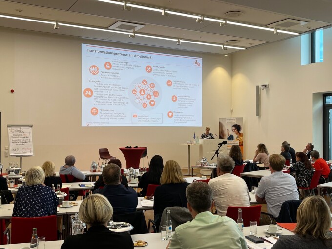 Viele Menschen sitzen an Tischen. Im Hintergrund eine Präsentationsfolie und am Rednerpult Ulrike Harstick-Tacke von der Regionaldirektion Berlin-Brandenburg der Bundesagentur für Arbeit (BA).