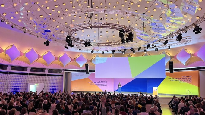 Totale über einen voll besetzten Konferenzsaal, im Hintergrund eine Rednerin auf einer Bühne.