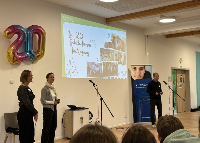 Mehrere Mitarbeitende der kobra.net gGmbH (u. a. Norbert Bothe ganz rechts) stehen vor einer virtuellen Leinwand mit Fotos früherer Veranstaltungen, an der Wand hängt ein Luftballon mit der Zahl 20.