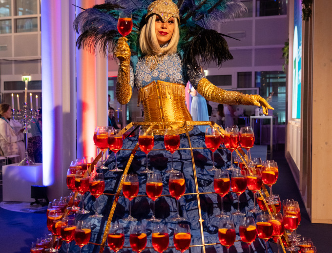 Eine Frau mit prachtvoller Robe und ein darüberliegendes Gestell, in dem gefüllte Weingläser stehen. Ein Glas hält sie zuprostend in der Hand.