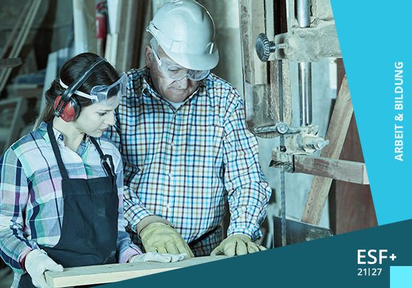Rubrik-Arbeit-und-Bildung (AdobeStock: Robert Kneschke) Eine junge Frau in Arbeitskleidung mit Gehörschutz und Schutzbrille wird von einem älteren Mann mit Helm und Schutzbrille an einem Bohrer angeleitet.